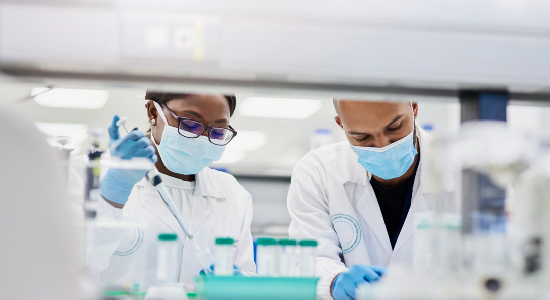 masked researchers in lab coats work with liquids at a lab bench.